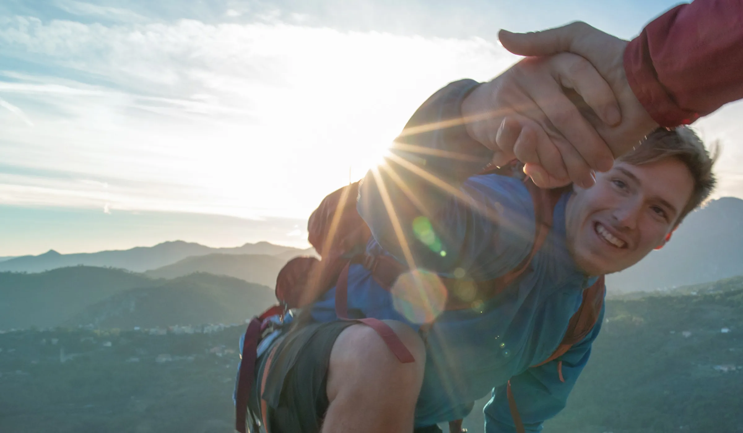 Ein Mann erklimmt einen Berg und greift nach der ausgestreckten Hand, die ihm oben entgegengestreckt wird. Die Szene vermittelt Durchhaltevermögen, Unterstützung und den Erfolg persönlicher Herausforderungen.