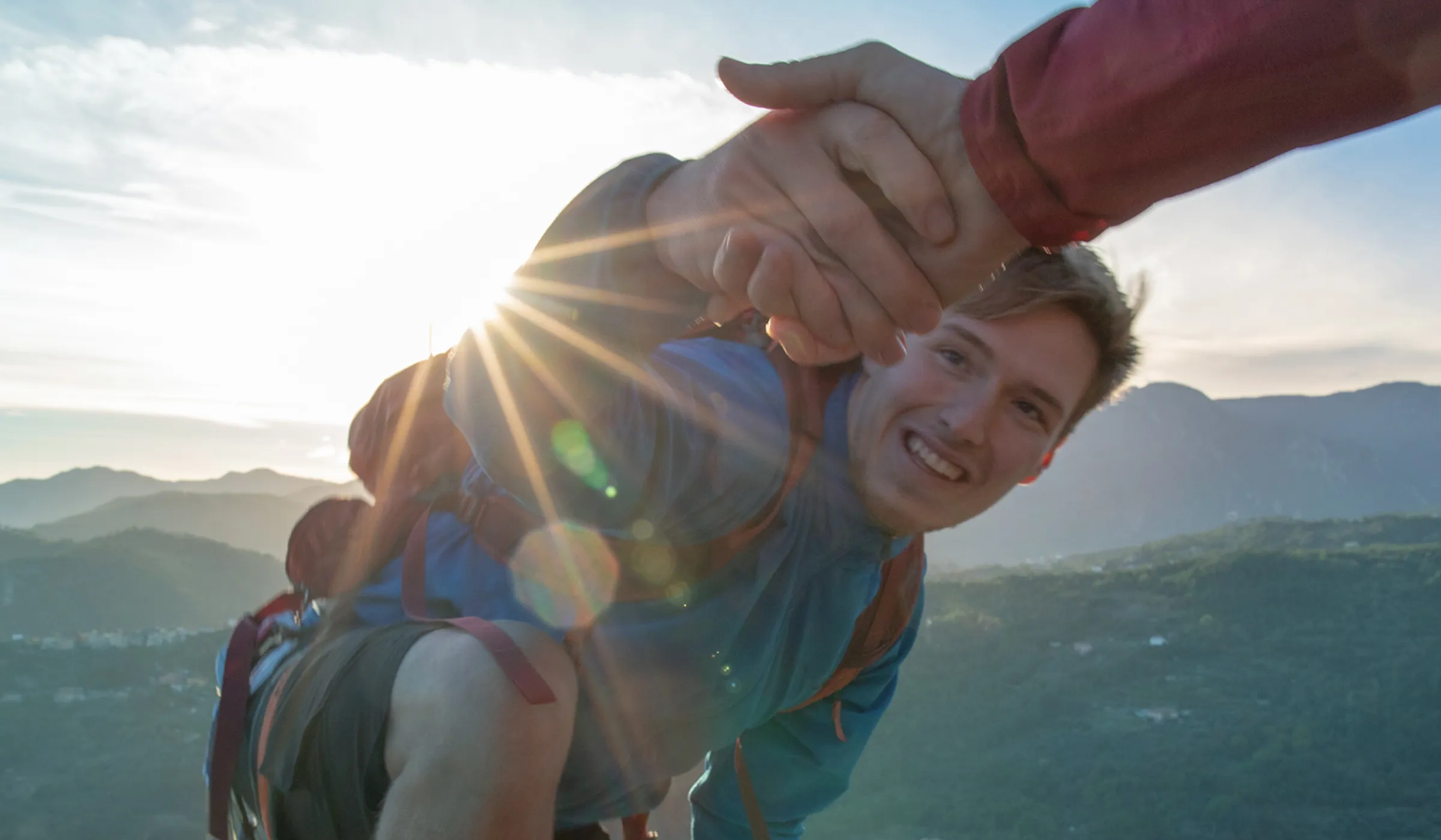 Ein Mann erklimmt einen Berg und greift nach der ausgestreckten Hand, die ihm oben entgegengestreckt wird. Die Szene vermittelt Durchhaltevermögen, Unterstützung und den Erfolg persönlicher Herausforderungen.