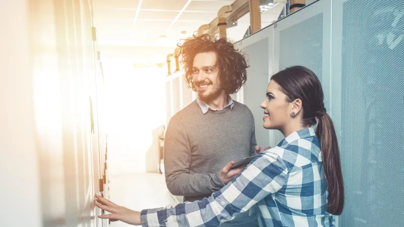 Ein Brainguards Mitarbeiter und eine Mitarbeiterin stehen vor einem Serverschrank und schauen freundlich, wobei der Mitarbeiter ein Tablet in der Hand hält. Die Szene vermittelt eine positive und kooperative Atmosphäre im Kontext der Servertechnologie.