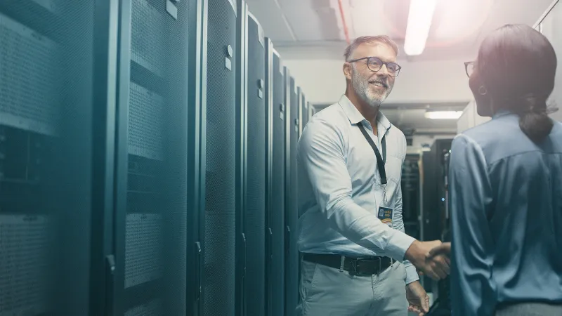  Ein Mann und eine Frau stehen zwischen Serverschränken und geben sich die Hand. Der Handschlag symbolisiert Zusammenarbeit und Partnerschaft im technologischen Umfeld.