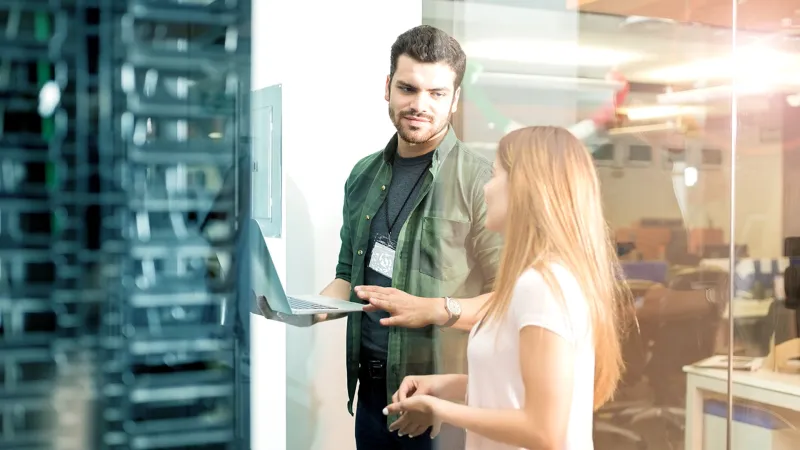 Ein Mann und eine Frau stehen vor einem Serverschrank in einem Büro und tauschen Blicke aus. Der Mann hält dabei einen Laptop in der Hand. Die Szene vermittelt einen Eindruck von Zusammenarbeit und technischer Inspektion innerhalb des Bürosettings.